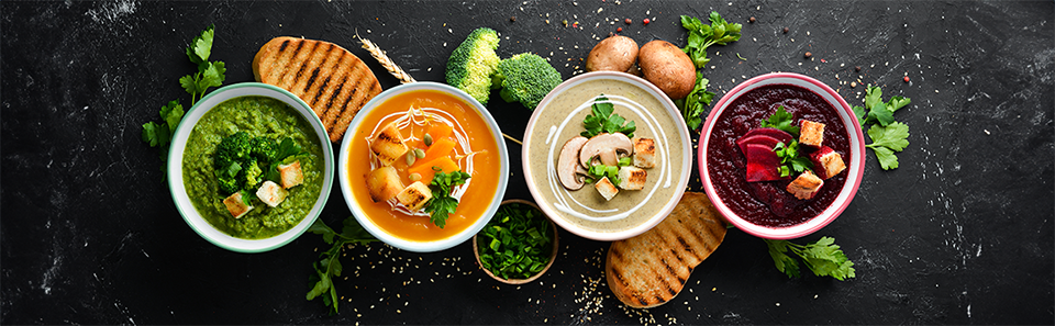 Vier kleurrijke kommen soepen (broccoli, groente, champignon en biet) voor het soepdieet met geroosterd brood en verse kruiden op een donkere achtergrond.